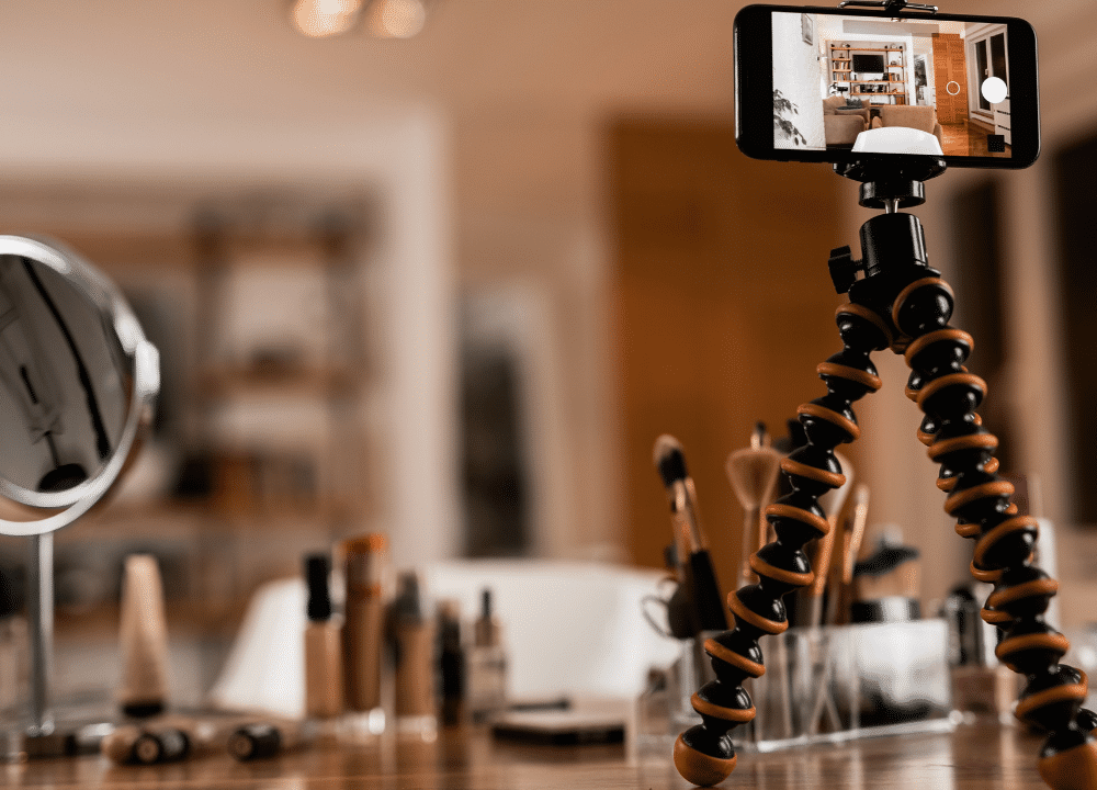 A smartphone on a tripod records a compelling video in an organized room with a desk full of makeup products, brushes, and a round mirror. Perfect for creating content that will capture attention in 2024.