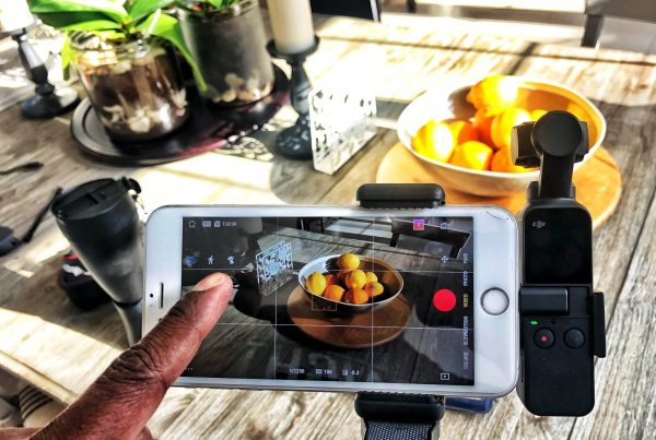 A person is using a smartphone mounted on a tripod to photograph a bowl of oranges on a wooden table. The smartphone screen displays the camera interface, focusing on the bowl of oranges, capturing attention for producing compelling videos in 2024.