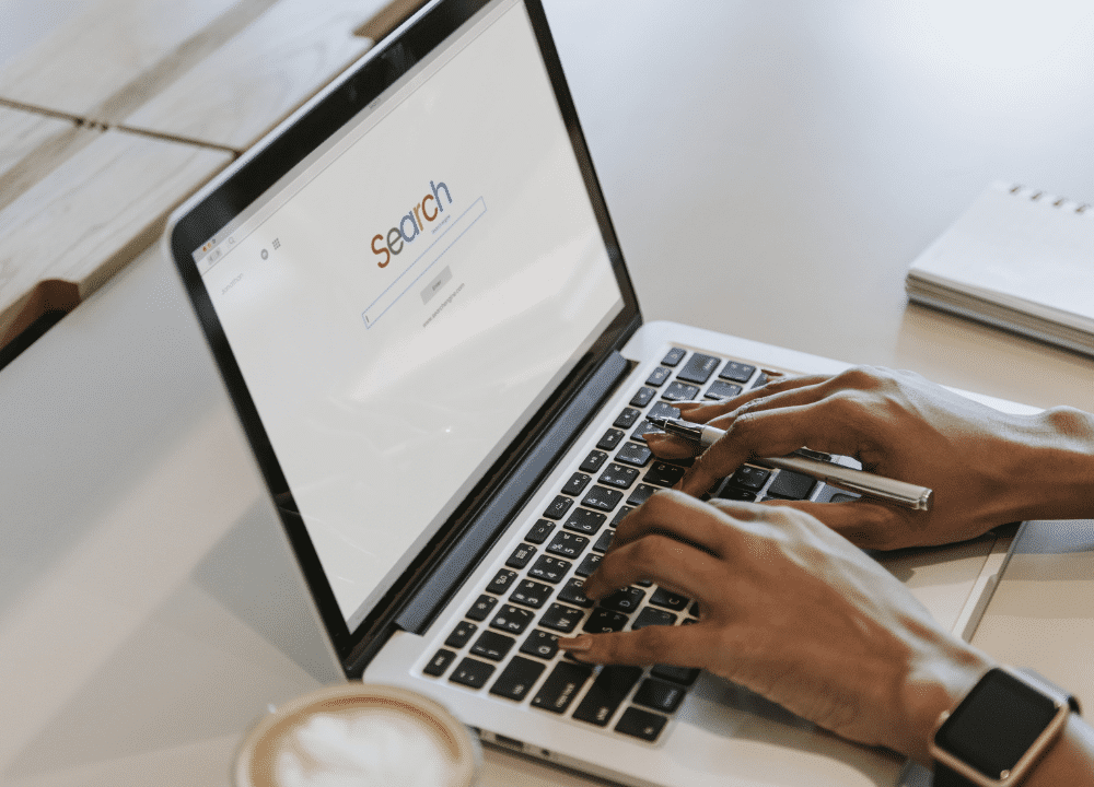 Person typing on a laptop with an SEO search engine screen open, a coffee cup in the foreground, and a notepad and pen nearby on the table.