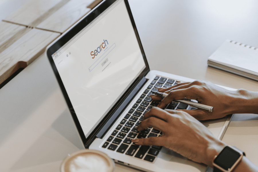 Person typing on a laptop with an SEO search engine screen open, a coffee cup in the foreground, and a notepad and pen nearby on the table.